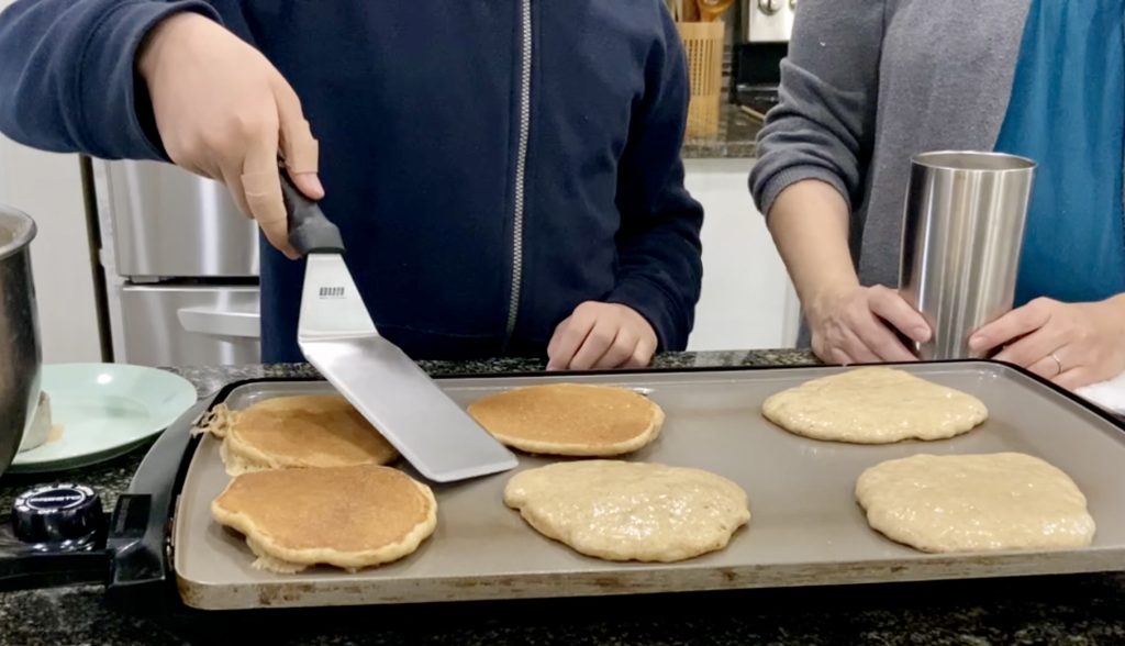 Child flipping overnight sourdough pancakes with mother standing nearby supervising 